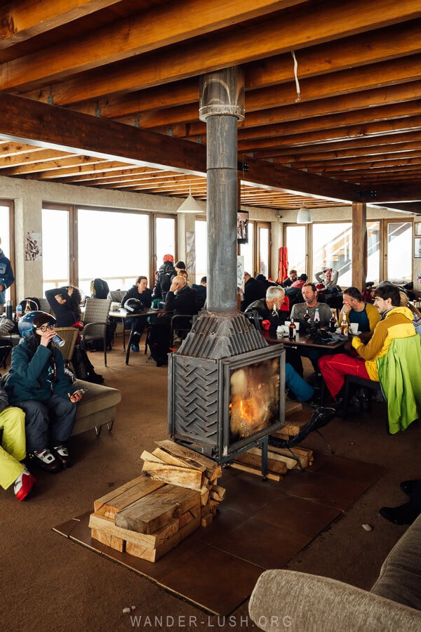 Cosy interior of Cafe Zuruldi in Mestia, Georgia, featuring a wood-burning stove at the centre, with skiers and snowboarders relaxing and enjoying meals after a day on the slopes.