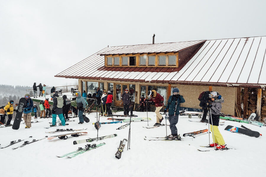 Skiers and snowboarders gathered outside Cafe Zuruldi in Mestia, Georgia, with ski equipment scattered in the snow on a cloudy winter day.