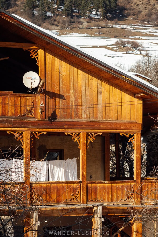 A traditional wooden balcony in Mestia, Georgia, adorned with intricate carvings, a satellite dish, and white laundry drying in the winter sun.