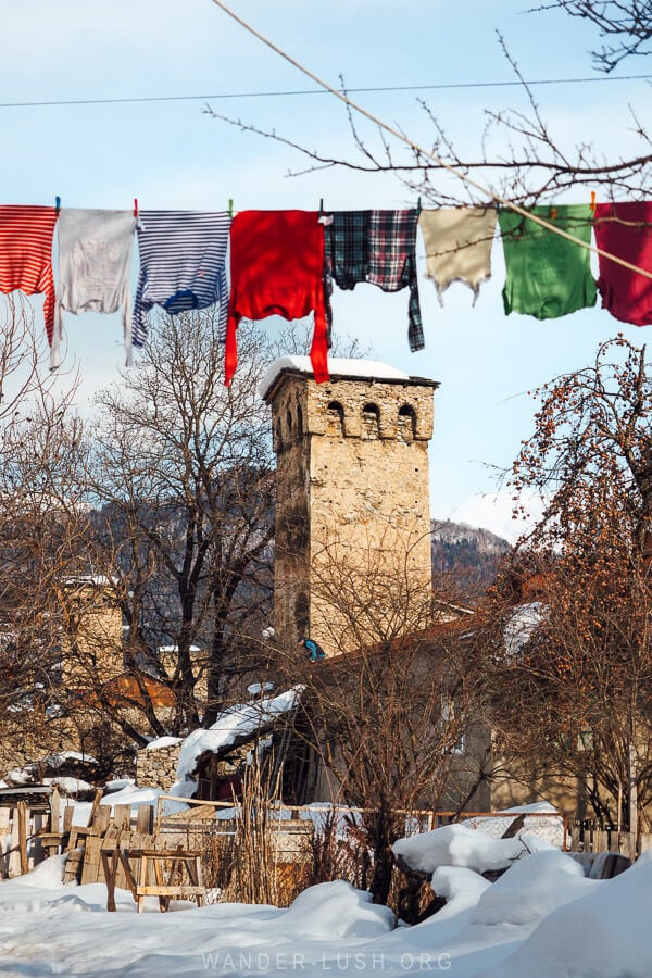 Colourful laundry hanging on a washing line in Mestia, Georgia, with a traditional Svan tower and snow-covered rooftops in the background.