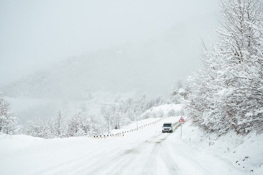 A car drives along a snow-covered mountain road towards Mestia, Georgia, surrounded by frost-laden trees and misty winter landscapes.