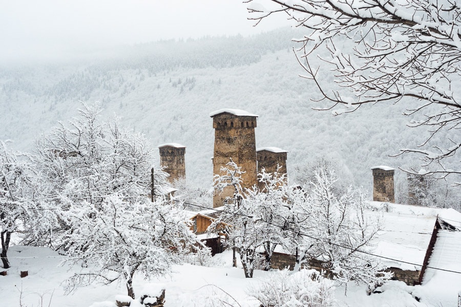 Snow-covered Svan towers in Mestia, Georgia, framed by frost-laden trees with misty mountains in the background.