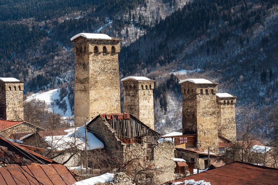 A cluster of ancient Svan towers in Mestia, Georgia, standing among rustic buildings with a backdrop of snow-covered mountains and dense forests.