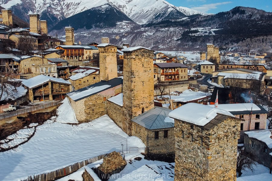 Aerial view of Ushba Street in Mestia, Georgia, showcasing multiple ancient Svan towers, stone houses, and a snowy mountain landscape in the background.