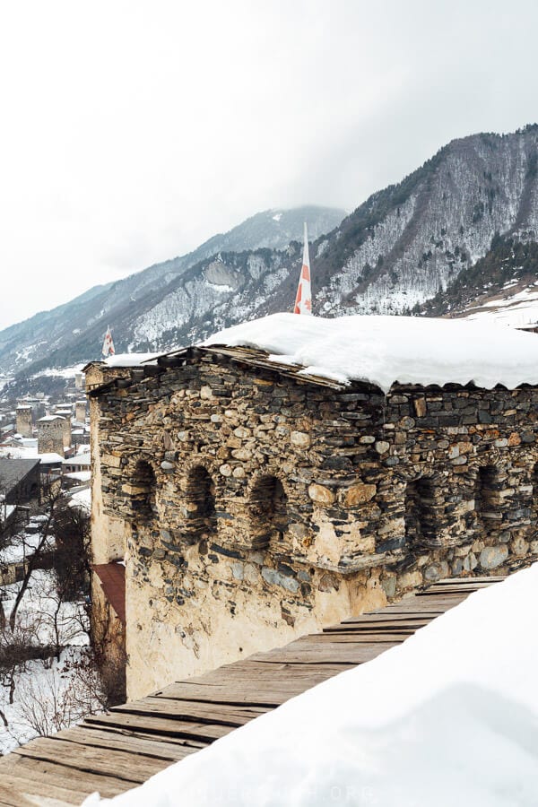 A snow-covered stone rooftop at the Ratiani House Museum in Mestia, Georgia, with arched openings, wooden walkways, and Georgian flags, overlooking the winter landscape.