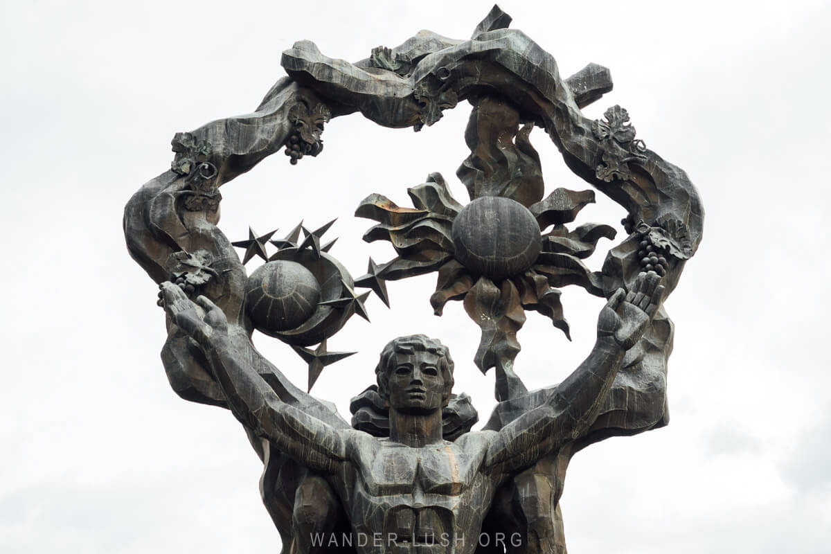 Bronze statue featuring a muscular figure with outstretched arms, holding a circular structure adorned with the sun, moon, and symbolic elements, set against a cloudy sky in Khobi, Georgia.