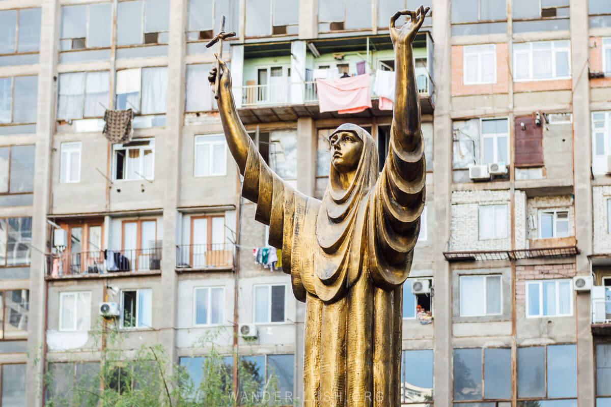 A golden statue of St Nino holding her downward turned cross, seen from inside a cemetery in Tbilisi against a backdrop of Soviet-era apartments.