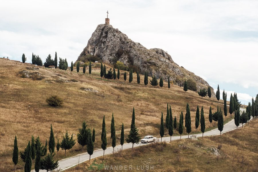A monastery sits atop a hill in autumn in Kakheti, with a tree-lined road winding below and a Lada car ambling along.