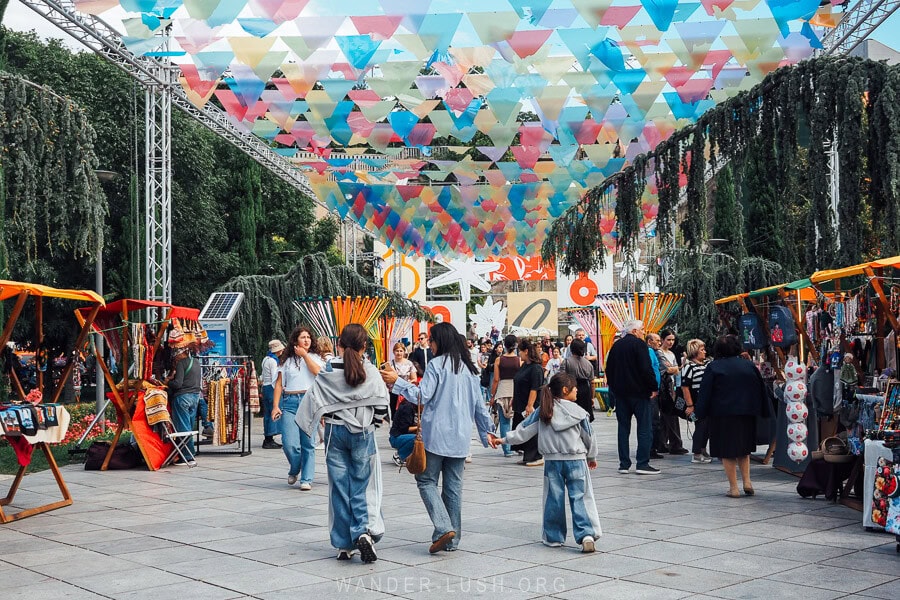 Young people walk beneath a colourful flag display alongside market stalls in Rike Park as part of the Tbilisoba autumn festival in Georgia.