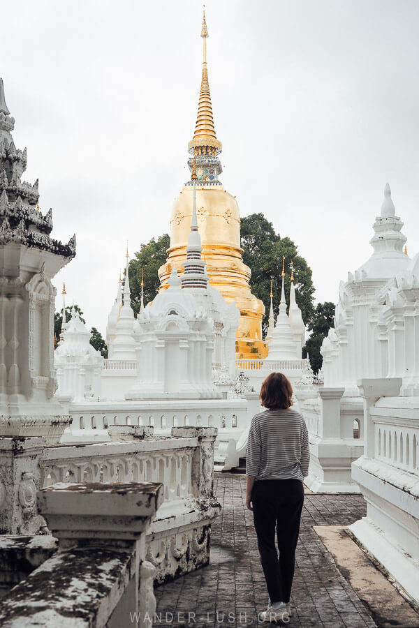 A woman walks through a temple complex with white stupas and a golden spire in Chiang Mai, Thailand.