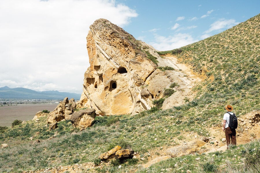 A man dressed in a hat looks up at a rock formation with burial chambers cut from its surface at the Kaspi Necropolis in Georgia.