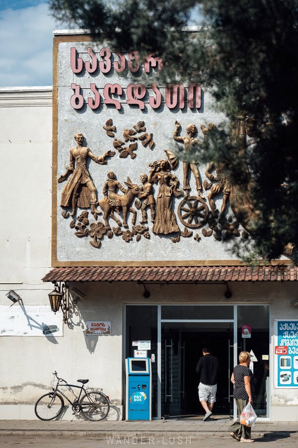 People walk along the sidewalk in Baghdati, Georgia, with an impressive Soviet-era relief sculpture on the facade of the former shopping centre.