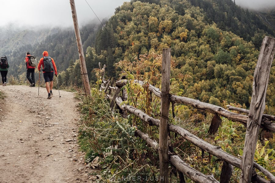 People walk into the distance along a dirt path next to a forested mountain in Svaneti.