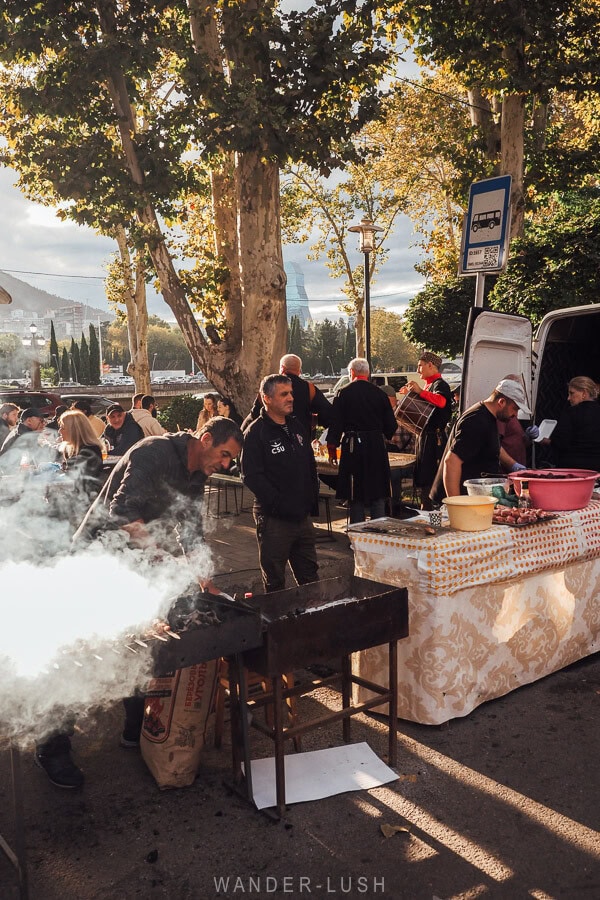 Men grill meat on portable BBQs at the Tbilisoba festival, with Tbilisi's skyline in the background.