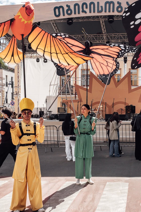 Street actors with birds and butterflies prepare for the Tbilisoba opening parade in front of a stage on Orbeliani Square.