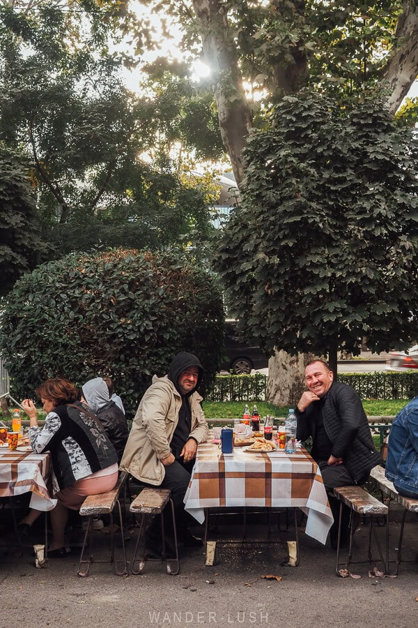 Two men seated at a table at Tbilisoba on the side of the street pose for a photo.