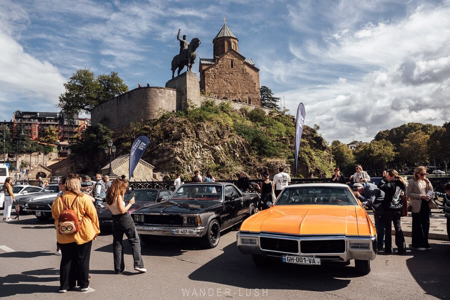 A vintage orange car parked on Metekhi Bridge in Tbilisi, with the Virgin Church and state in the background and the bridge closed to traffic during Tbilisoba festival.